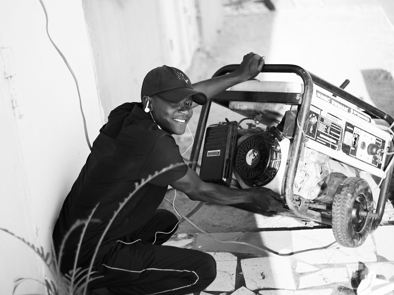 A cheerful young man wearing a cap and earphones operating a portable generator outside.